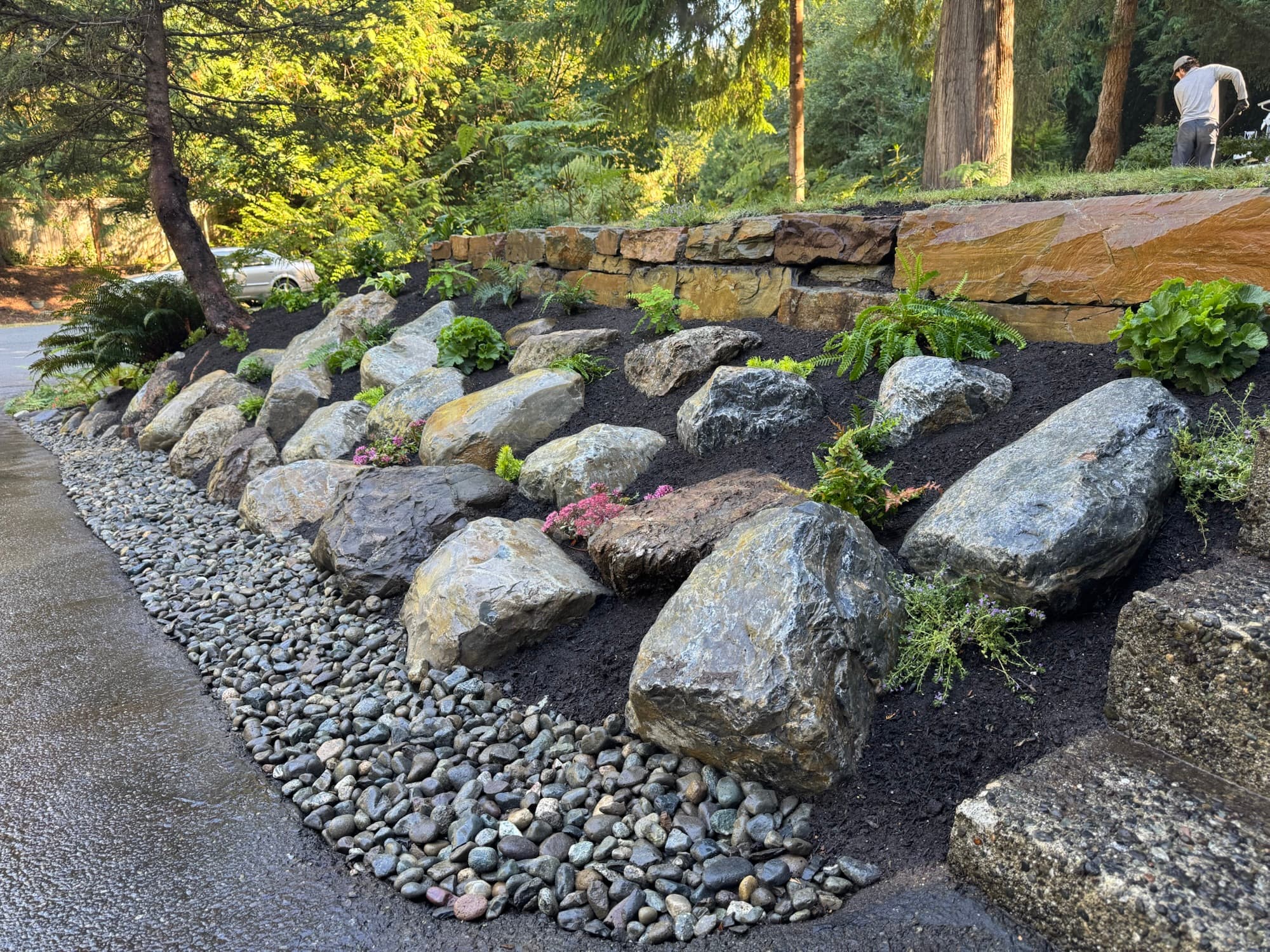 A tiered boulder retaining wall set into dark mulch with river-rock drainage and new planting along a drive.