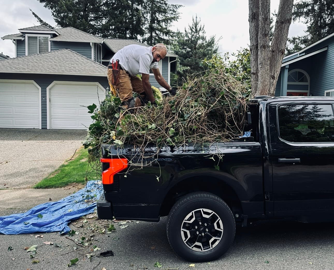 An electric pickup loaded with cleared brush and salvaged plant material at the kerb of a suburban home.